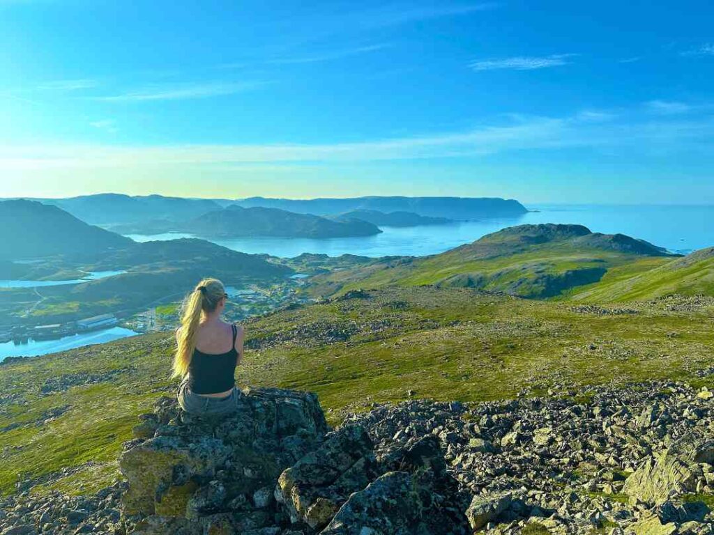 A woman in a thin summer top sitting on a rock on the top of a mountain during summer, with berathtaking views over the greenery and fjords below under a deep blue sky