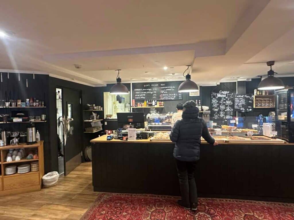 A cafe counter with lots of pastries, a black wall with hand written menus, and a guest waiting at the counter standing on the red rug on the wooden floor, warm lighting. 