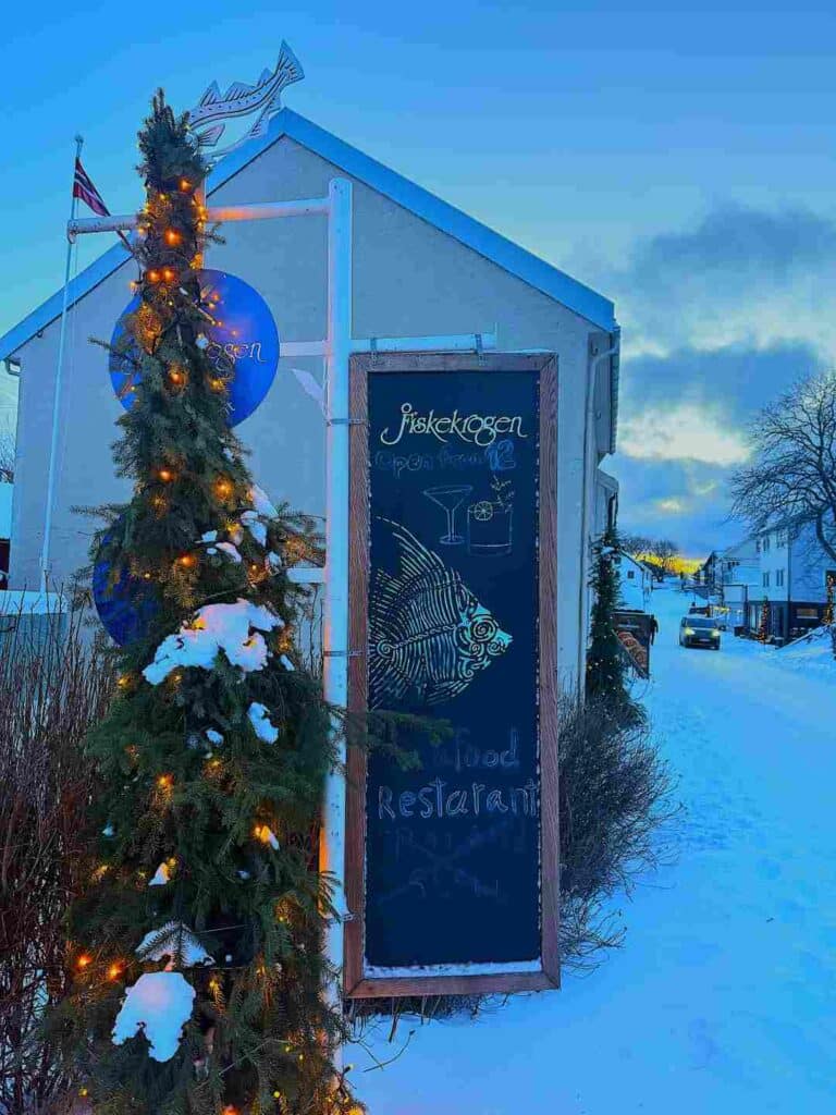 A snow covered street with a green bush with twinkeling lights next to a hand writter restaurant sign, in Henningsvær, Lofoten in blue winter daylight 