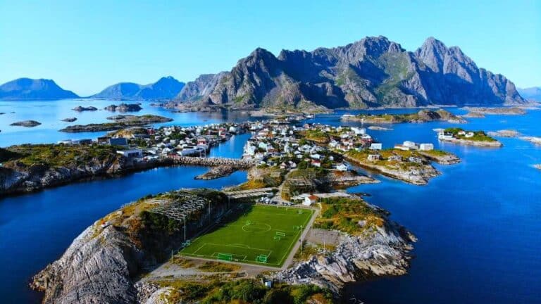The famous green football field on Henningsvær in Lofoten Norway, from the air. The green field covering almost the entire island, and historic wooden white and red houses scattered on the small islands around, surrounded by deep blue sea under a clear blue summer sky, majestic mountains in the distance.