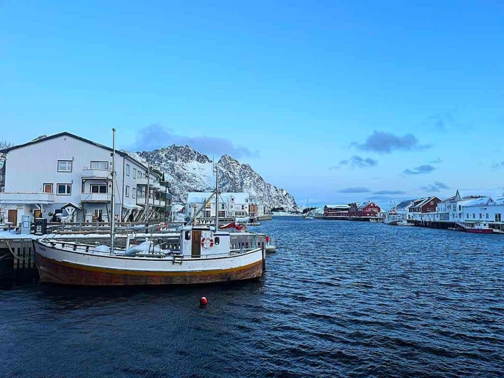 A dark harbor in Henningsvær, Lofoten in Norway in winter, surrounded by old wooden houses, with a wooden fishing boat at the dock under a blue winter sky