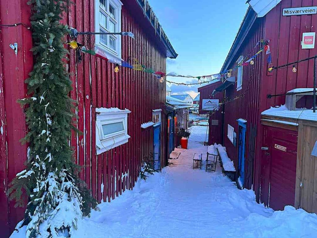 A narrow alleyway between old red wooden houses on a winter day in Henningsvær Lofoten in Norway, snow covering the ground, and you can see the pale blue sky over the alleyway that is decorated with small colorful flags