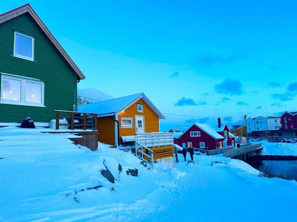 A snowy winter path by a harbor, surrounded by colorful wooden houses, lots of snow, under a pale blue winter sky in Lofoten Norway