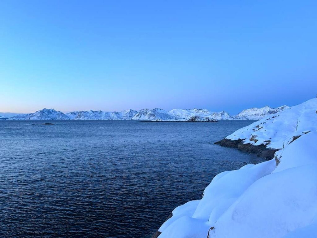 Dark blue water surrounded by snowy shores, across the vast fjord are white snow covered mountain ranges of the Lofoten Islands in the blue winter light in Norway