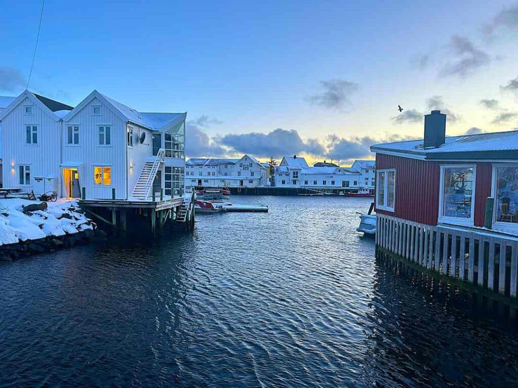 A small harbor with dark waters surrounded by red and white old wooden houses on a winter day in Lofoten Norway under the blue winter light, and a pale blue sky