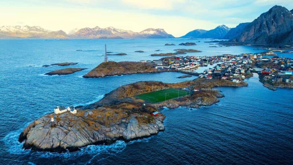 A deep green football field covering almost an entire island, short of a cluster of white wooden buildings at the tip of the island, on the dramatic edge of Henningsvær in Lofoten, surrounded by dark blue Arctic waters and mountain ranges under a pale blues sky
