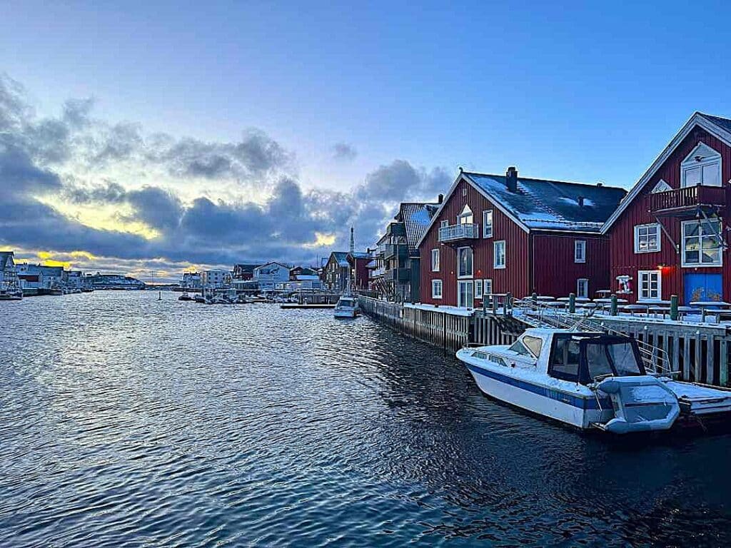 Harbor area in Henningsvær, Lofoten, during winter. The harbor is lined with red wooden houses by a wooden pier, boats on the water, undera clear blue winter sky