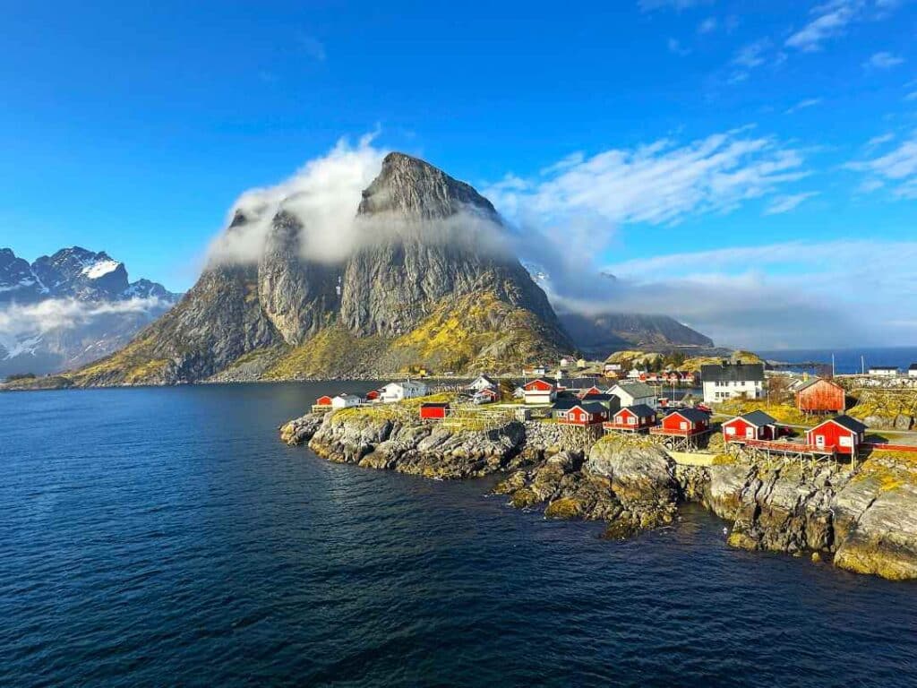 Red wooden fishermans cabins dotted all over an island, surrounded by dark blue waters, and mountains towering in the distance partly covered by almost transparent floating clouds under a clear blue sky