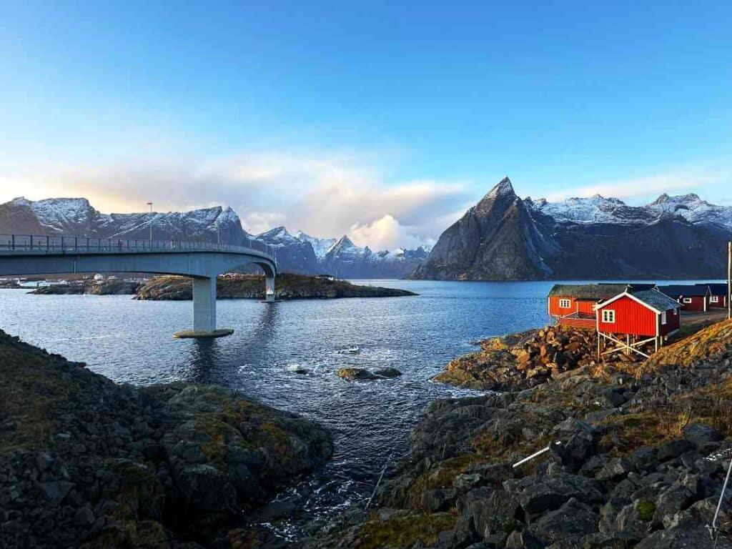 A row of red wooden fishermans cabin lined by the shore of a fjord, a majestic bridge connectiing the next island across the fjord, and steep mountain peaks covered in snow in the distance under a blue sky