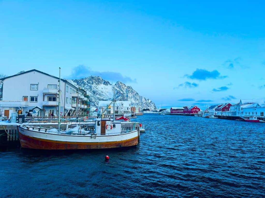 A wooden fishing boat at the dock next to white and red wooden houses under a rugged mountain and a soft blue sky
