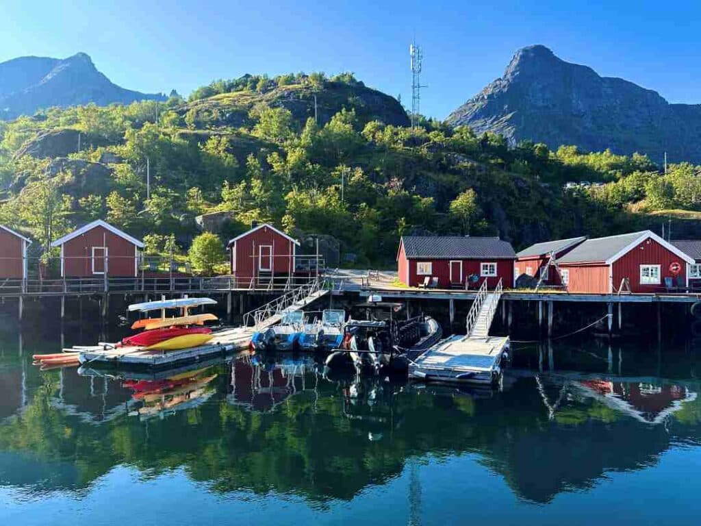A tiny rorbu area by the docks, with kayaks stacked on the jetty, under a green hill, with steep mountains in the distance under the clear blue summer sky