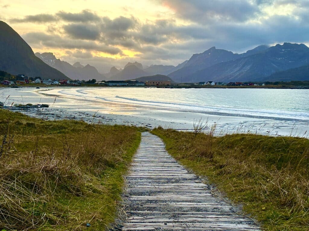 A wooden pathway heading downhill to a white sandy beach, a silver sea surface with soft waves rolling in, and in the distance majestic mountain ranges into the distance under a golden sky during sunset in Lofoten, Norway