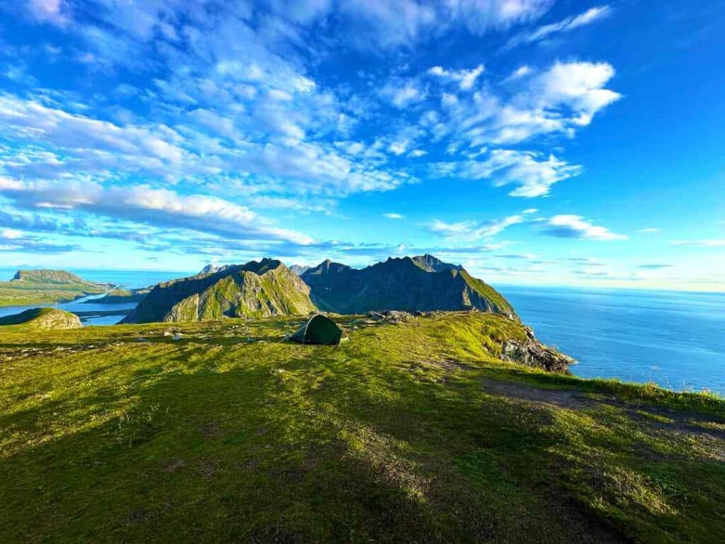 A bright green mountain plain with a small tent, with view to several mountains in the distance, before the deep blue sea stretches indefinitely under a clear blue sky in the summer in the Lofoten Islands Norway