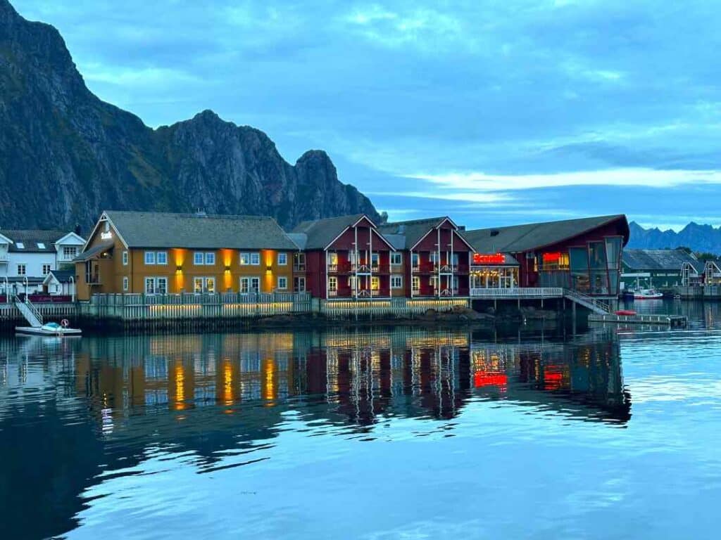 Red and yellow wooden houses by the shore of a fjord, under a dusky blue evening sky, under magnificent dark grey mountains looming in the background