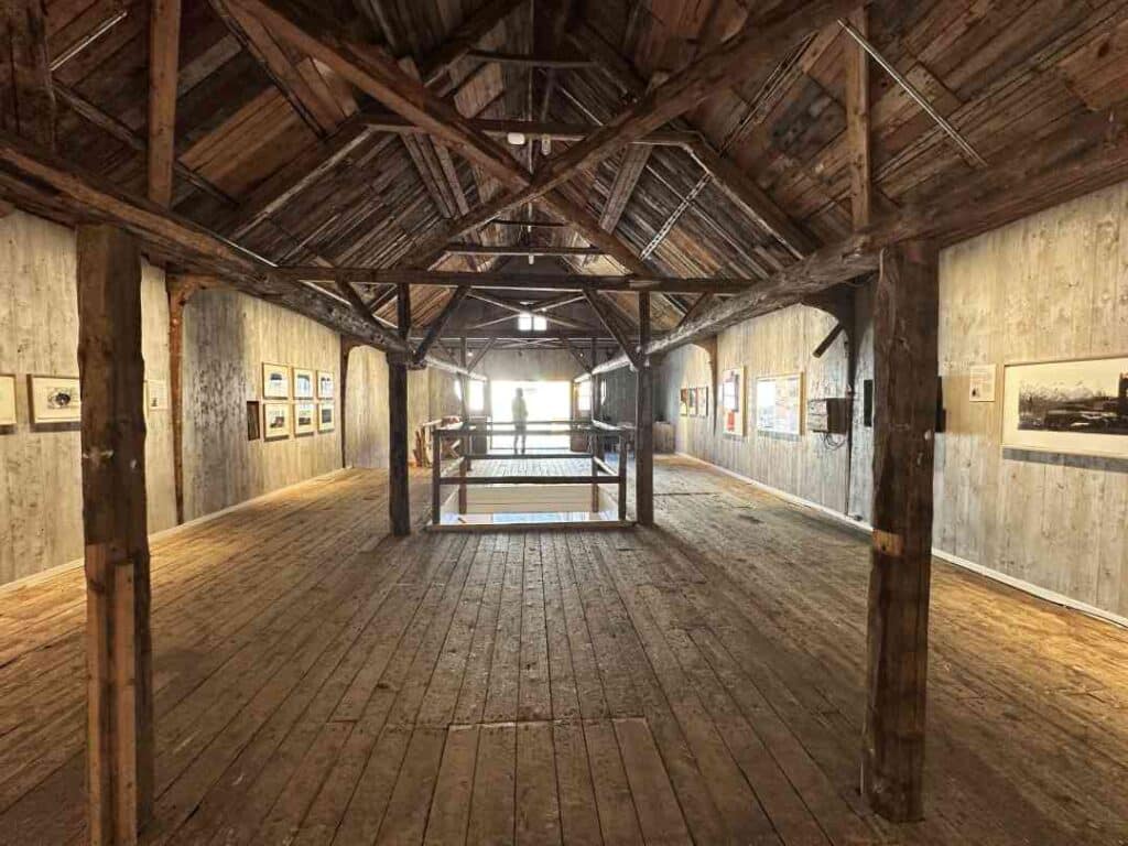 A spacious airy art gallery with old fashioned natural woodwork and columns, and grey walls with art displayed in natural light from the far side windows