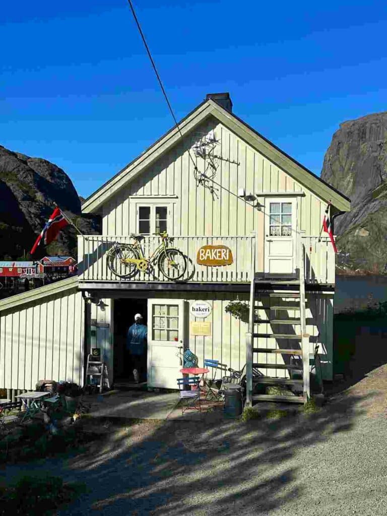 A white wooden charming old house with a bakery sign, decoreated with ornaments, nestled between mountains and bathed in sunlight under a clear blue sky