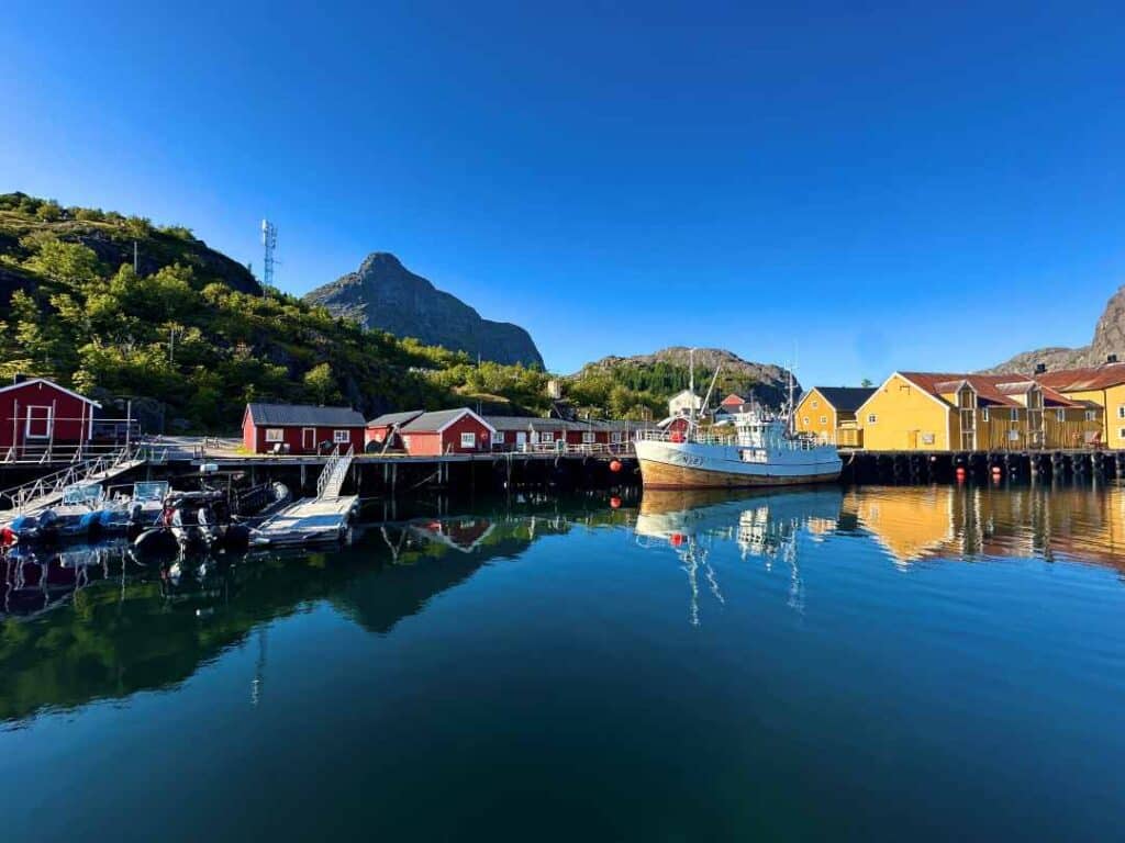 A wooden fishing boat by the dock floating on deep blue water surrounded by red and yellow wooden fishermans cabins under a green hill and a deep blue sky during summer in Nusfjord, Lofoten, Norway