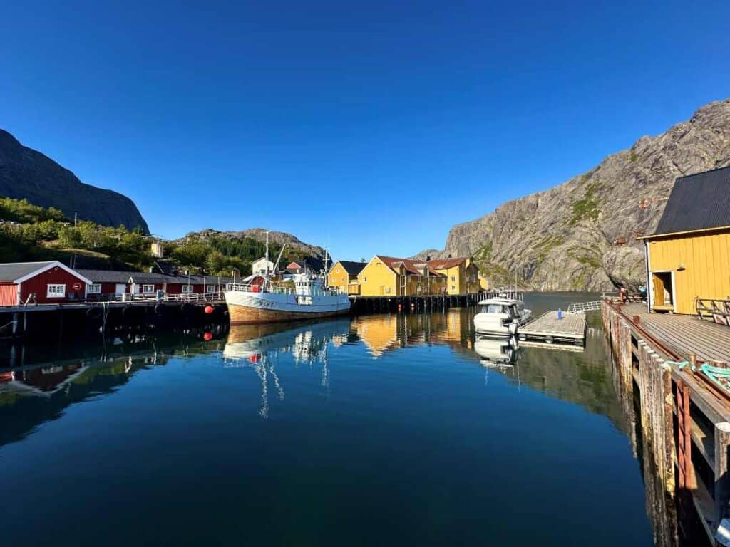 The harbor of a traditional fishing village, surrounded by yellow and red wooden fishermans cabins, tucket inside grey rocky mountains under a blue summer sky