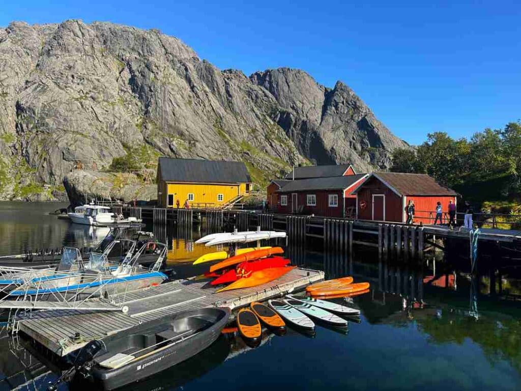 Kayaks and speed boats ready to go on a floating jetty surrounded by red and yellow fishermans cabins on a bright sunny summer day under the mountains in Nusfjord, Lofoten, Norway