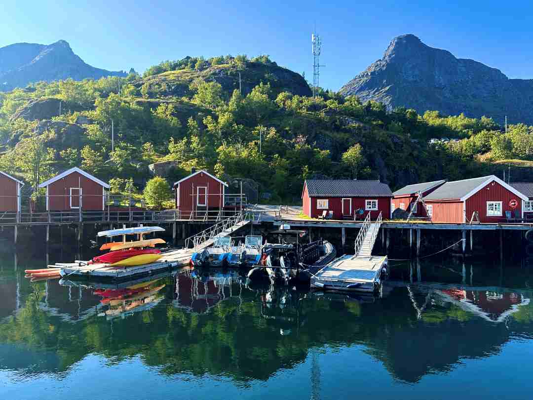 Red fishermans cabins tucked under a green hill during summer sitting on the shore of the deep blue fjord under a blue sky in Nusfjord, Lofoten, in summer