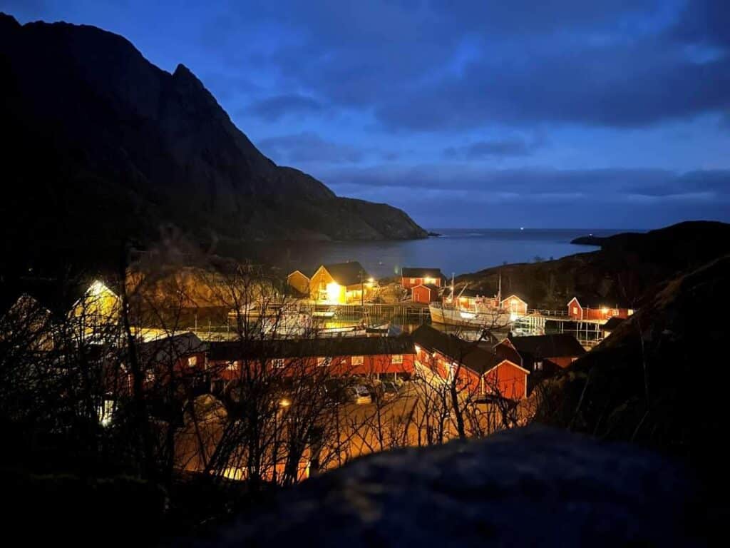 The small village Nusfjord seen from above at night, warm lights surrounding the red and yellow fishermans cottages, mountains steeping into the fjord in the distance under a dusty blue night sky in summer Lofoten, Norway