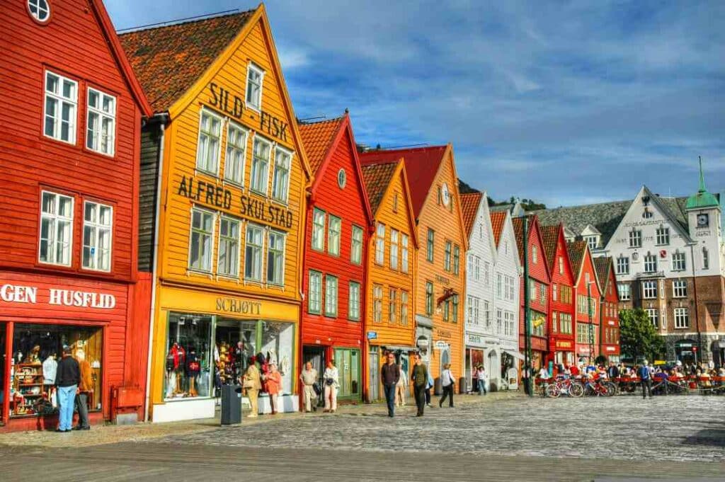 Colorful historic four storey wooden houses in red, orange, yellow and white under a blue sky, along a pier with lots of people walking the cobblestoned walkways, in the historic docks of Bergen city, Norway on a sunny summer day.
