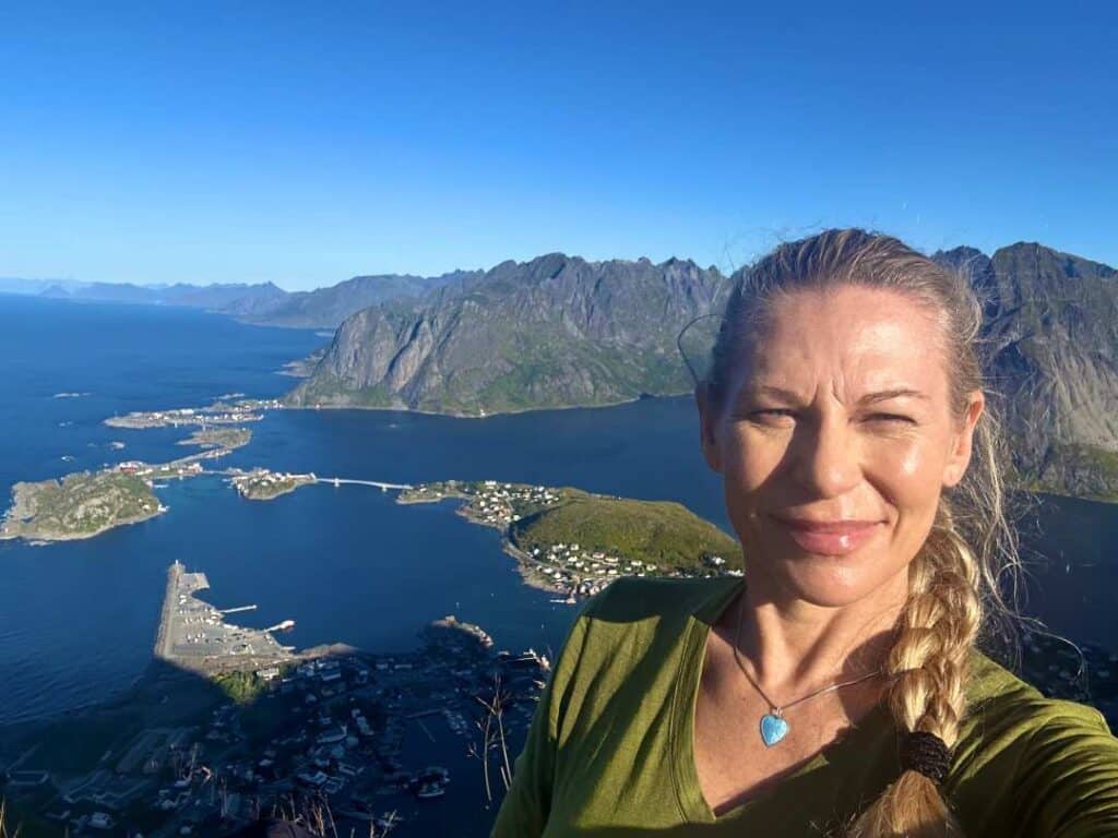 Blond lady smiling at the camera from the top of Reinebringen, Lofoten, on a clear sunny September day to a backdrop of Reine village far below, and the "Lofoten wall" mountains following the fjord line in the distance. 