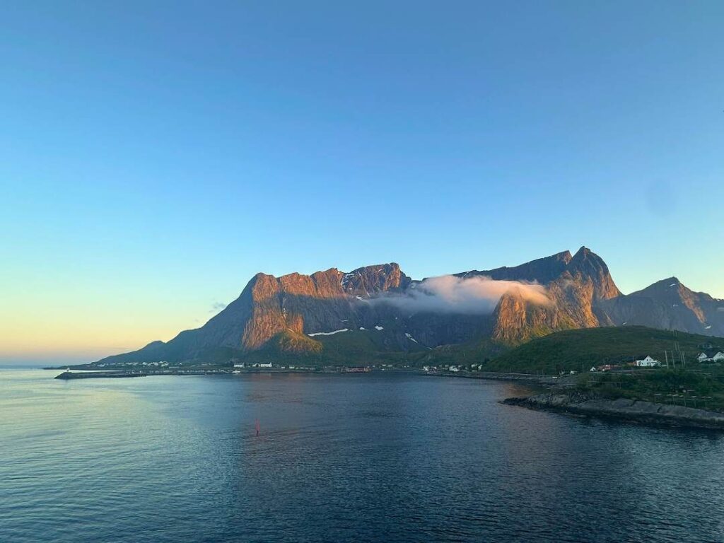 Reinebringen mountain ridge bathed in the glowing midnight sun under a clear blue sky at midnight in Reine Lofoten, surrounded by blank, calm deep blue seas