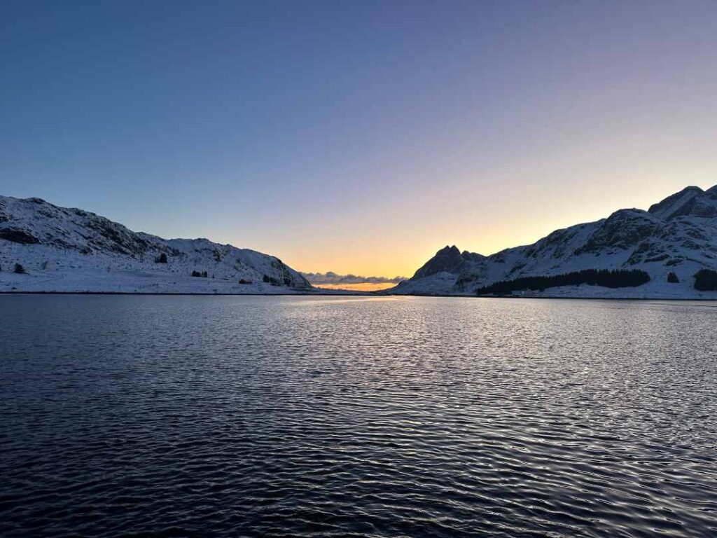 An almost blank winter fjord surrounded by snow covered mountains under a cold blue sky, with golden light on the horizon from the sun just below the horizon during the Polar Night in Lofoten Norway