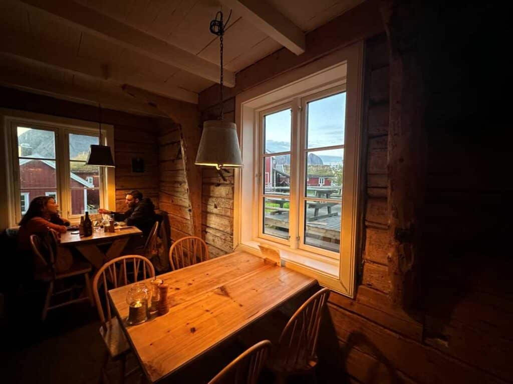 A rustic old wooden restaurant venue with warm lighting, original wooden walls, and view to red fishermans cabins and the fjord and mountains outside the windows in Lofoten Norway