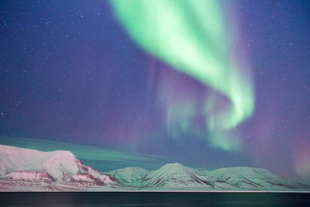 A light green and purple Northern Lights dancing in a starry sky over snow capped mountains across a dark cold sea in Svalbard, Norway