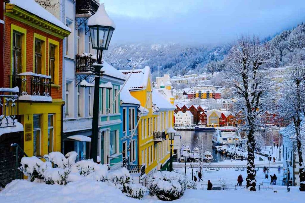 A steep street with colorful houses in blue, yellow and greens on a winter day, the ground and trees covered in snow, and below you see the harbor with boats and old wooden colorful houses. Winter in Bergen, Norway