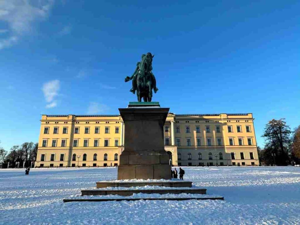 A large yellow building, the Royal castle in Norway, behind a large open plaza covered with snow under a blue sky, with a large statue in front of a historic figure mounted on a horse. Oslo, Norway in winter.
