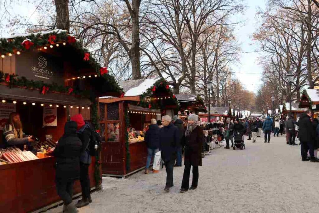 Small wooden cabin stalls in a Christmas market, selling food and Christmas decorations, along a snowy pedestrian road with lots of people strolling enjoying the vibe on a winter day in Oslo, Norway
