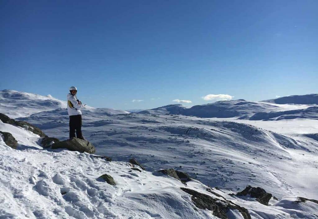 A skier in ski clothes wearing a helmet, on top of a snow covered mountain, with a backdrop of white snowy plains under a pale blue winter sky in Hemsedal, Norway