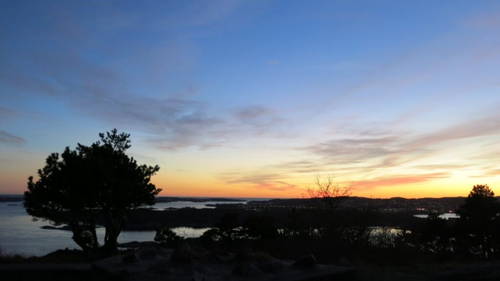 A glowing sunset under a pale blue sky, over low islands and the ocean behind small trees and bushes in Kristiansand Norway