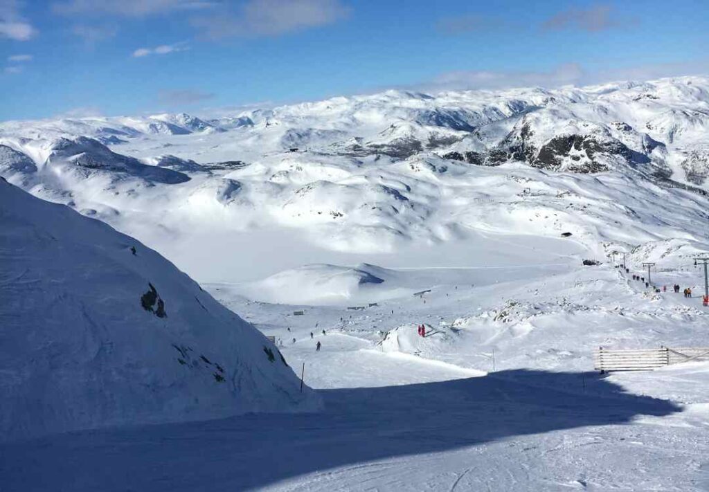 From the top of an alpine skiing resort, looking down on a neatly prepped slope, and beyond it are hundreds of snow covered mountain tops in the distance under a pale blue sky. Hemsedal, Norway in the winter.