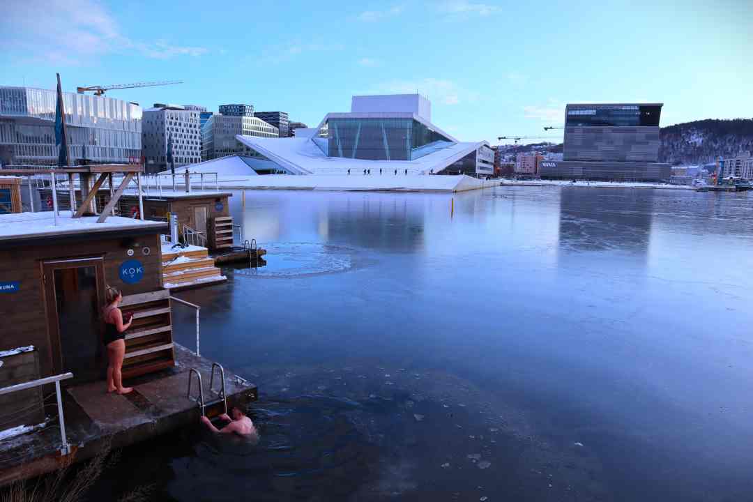 An ice covered fjord, with floating saunas on one side, and the Oslo Opera house on the other under a pale blue sky. Winter in Oslo Norway.