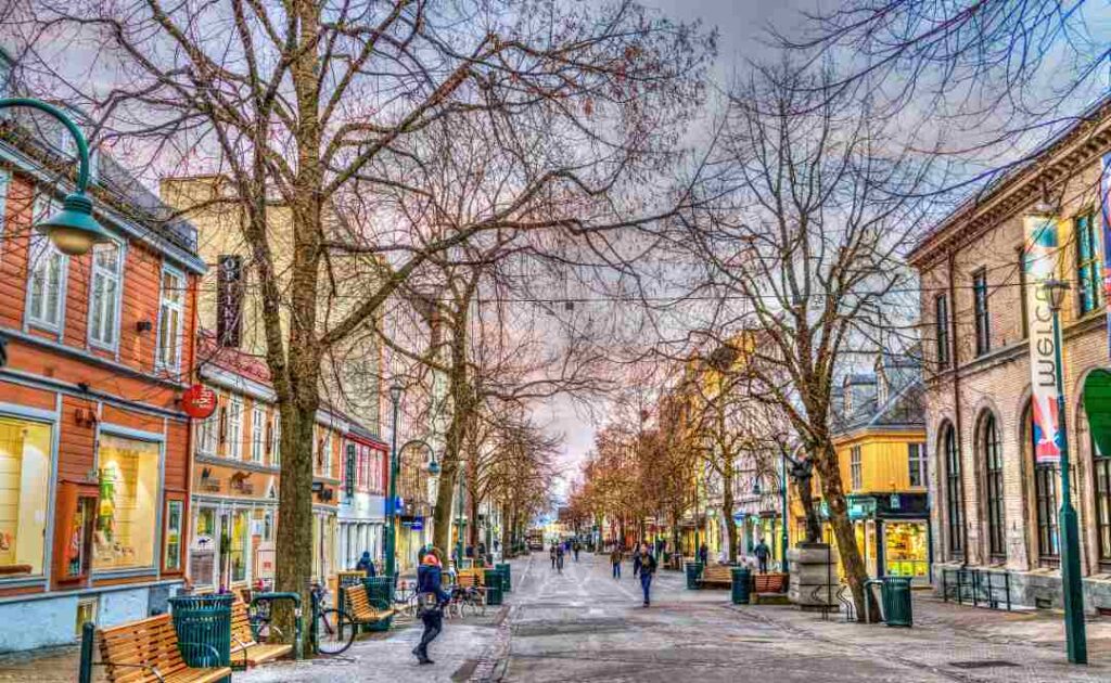 A main street with colorful old wooden houses on both sides, naked trees along the road under a pale pinkish sky, with people strolling. Winter in Trondheim Norway.