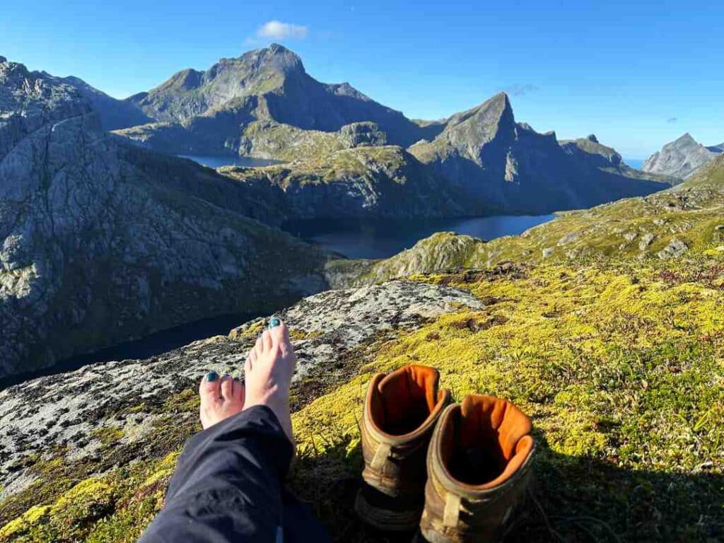 Hiker resting with boots off on the top of a mountain with views to a vast mountain range in the summer, green tops under a deep blue sky. Lofoten, Norway.