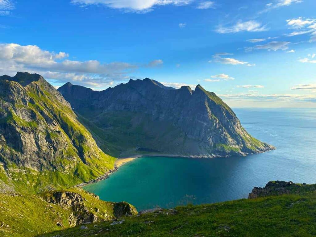 View from a mountain top towards other peaks and a secluded white sandy beach under green mountainsides beside emerald waters under a blue sky. Ryne, and Kvalvika Beach, Lofoten.
