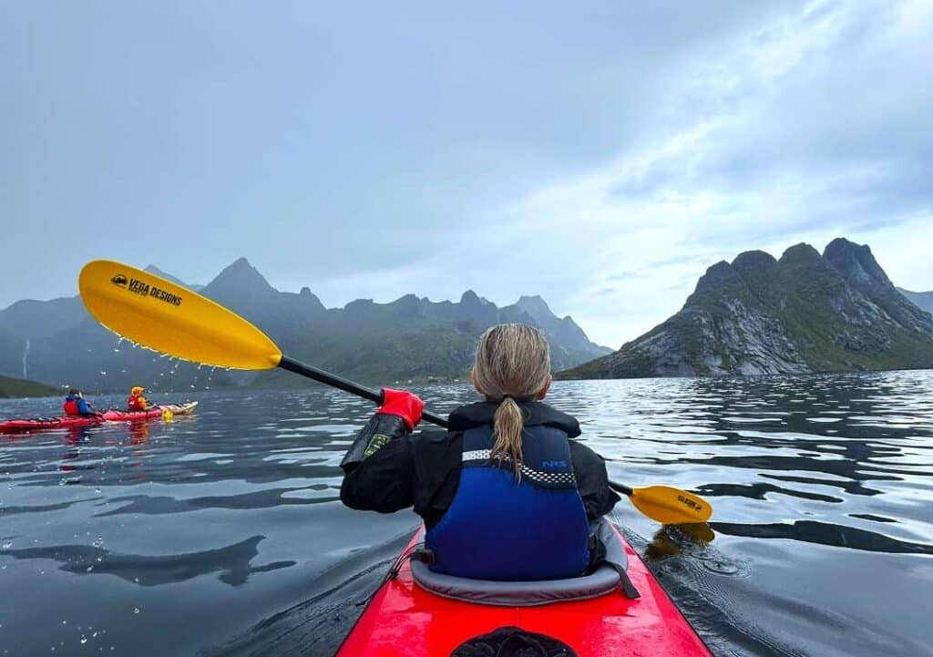 A blond woman kayaking under a dramatic sky in on a fjord surrounded my majestic mountains in the Reine Fjord, Lofoten, Norway.