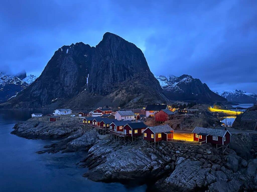Red fishermens cabins with glowing windows sitting on the edge of the fjord on the rocks. with a backdrop of a majestic mountain under a bluish sky at dusk in Lofoten Norway
