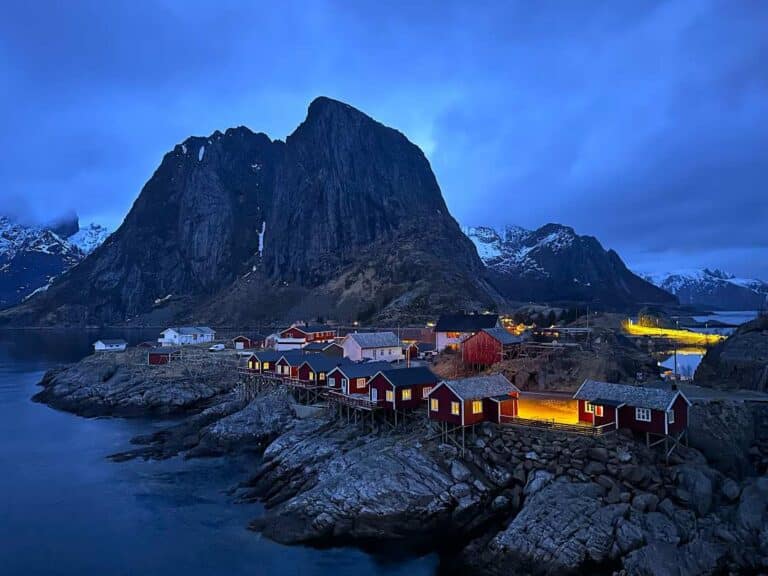 A small fishermans village with red cabins at dusk under a deep darkening blue sky, under a majestic mounatin hoovering in the distance