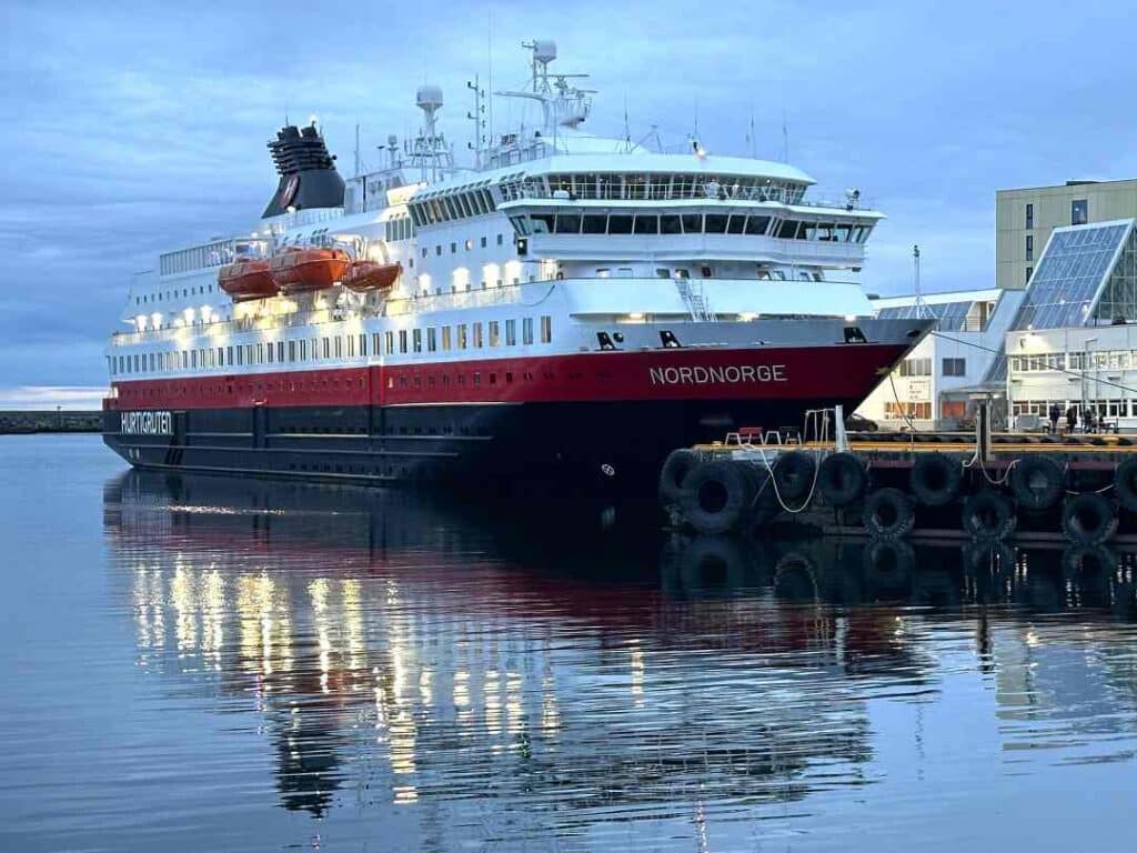 A large white, red and black vessel called Nordnorge, part of the Hurtigruten Coastal Voyage off the coast of Norway. Here docked in Svolvær in Lofoten on a late afternoon in September, it is dusk and a grey cloud cover over the sea and harbor.