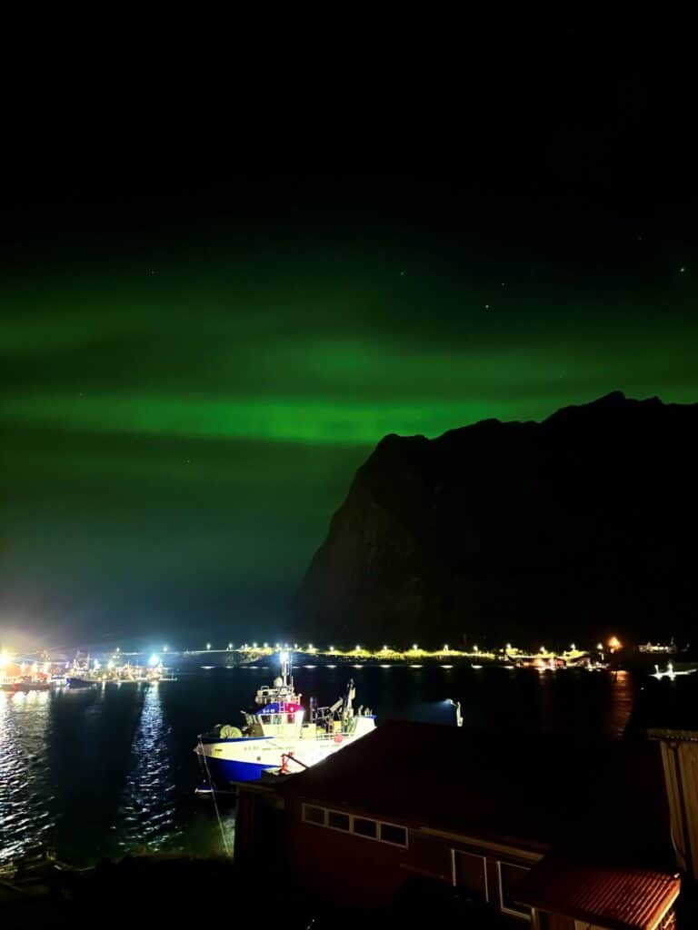 Faint green northern lights over Reinebringen mountain in Lofoten, with the Reine Fjord and fishing boats in the center on the dark waters.
