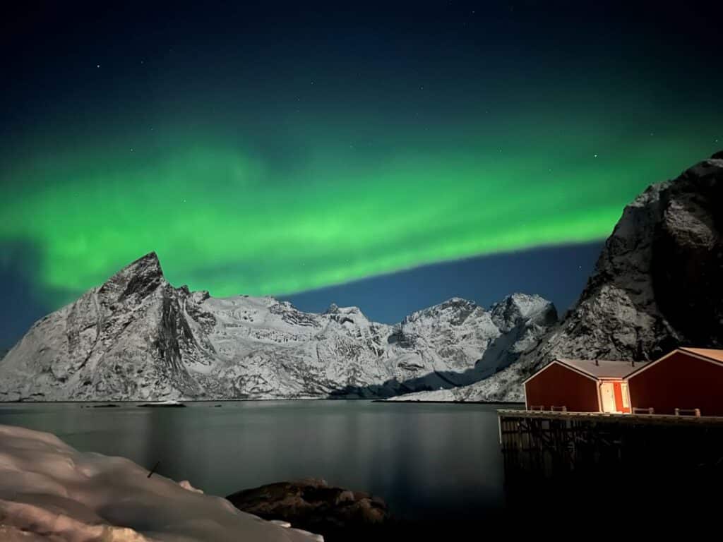 Green northern lights in a night sky over snow covered mountain across a fjord, with red fishermans cabins over the water.