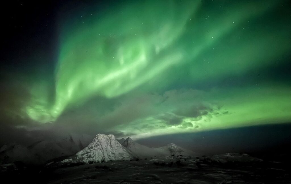 Green waves of Northern Lights dancing over a night sky over a snow capped mountain in Lofoten Norway in a clear winter night. 