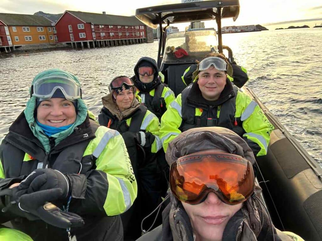 A happy group of people in yellow and black survival dresses, with goggles and hats on a RIB boat, ready for a RIB safari with the rd and yellow wooden houses of Svolvær Lofoten in the background.