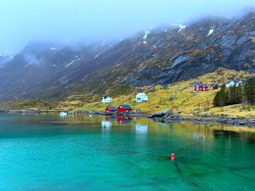 A handful of white and red fishermans homes on the yellow grass fjord shore under dark grey majestic mountains on a glooomy day with low clouds hovering hiding the mountain peaks. The fjord is blank and emerals, reflecting the houses on the shore. 